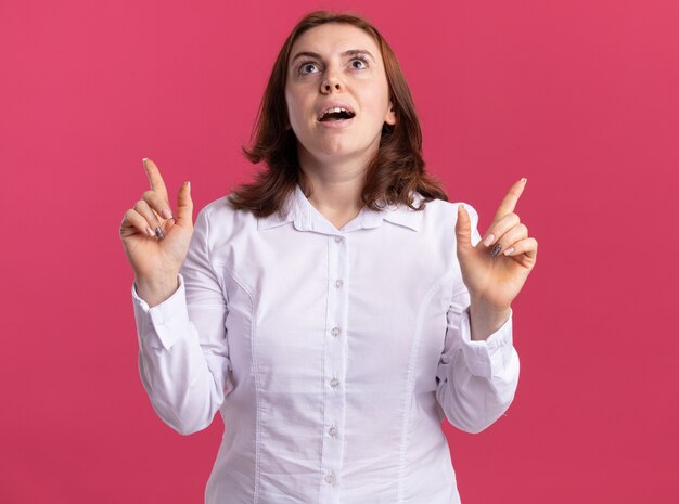 Mujer joven en camisa blanca feliz y emocionado apuntando con el dedo hacia arriba sonriendo de pie sobre la pared rosa