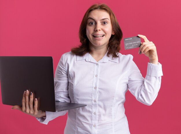 Mujer joven en camisa blanca con laptop mostrando tarjeta de crédito mirando al frente sonriendo alegremente de pie sobre la pared rosa