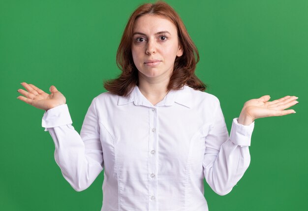 Mujer joven con camisa blanca mirando al frente confundido extendiendo los brazos hacia los lados de pie sobre la pared verde