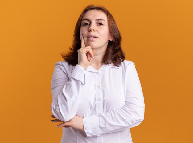 Mujer joven con camisa blanca mirando al frente con expresión pensativa pensando de pie sobre la pared naranja