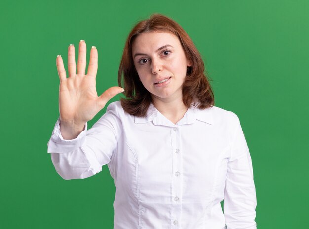 Mujer joven con camisa blanca mirando al frente mostrando y apuntando hacia arriba con los dedos número cinco sonriendo anding sobre pared verde