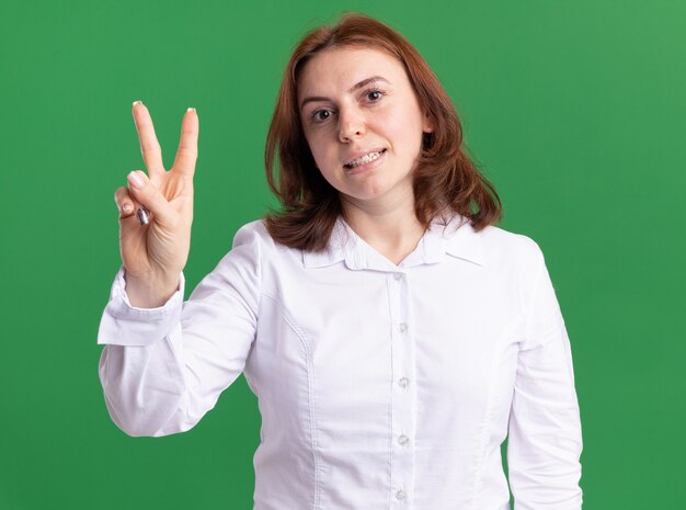 Mujer joven con camisa blanca mirando al frente mostrando y apuntando hacia arriba con los dedos número dos ying sobre pared verde