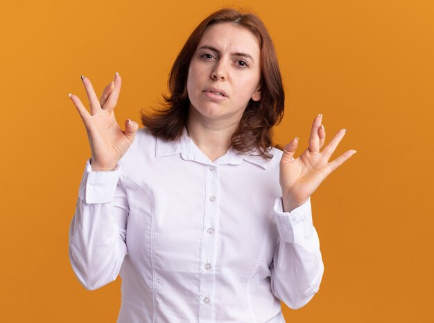 Mujer joven con camisa blanca mirando al frente con rostro serio cruzando los dedos de pie sobre la pared naranja