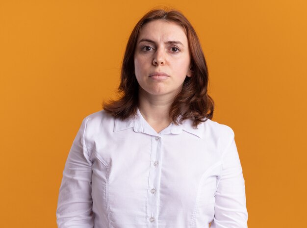 Mujer joven con camisa blanca mirando al frente con seria expresión de confianza de pie sobre la pared naranja