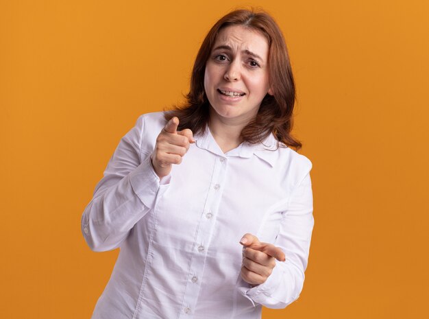 Mujer joven con camisa blanca mirando al frente sonriendo apuntando con los dedos índices en la parte delantera de pie sobre la pared naranja