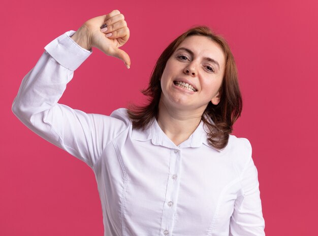 Mujer joven con camisa blanca mirando al frente sonriendo confiada apuntando a sí misma con el pulgar de pie sobre la pared rosa