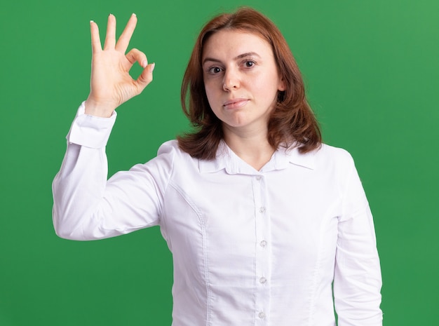 Mujer joven con camisa blanca mirando al frente sonriendo mostrando signo ok de pie sobre la pared verde