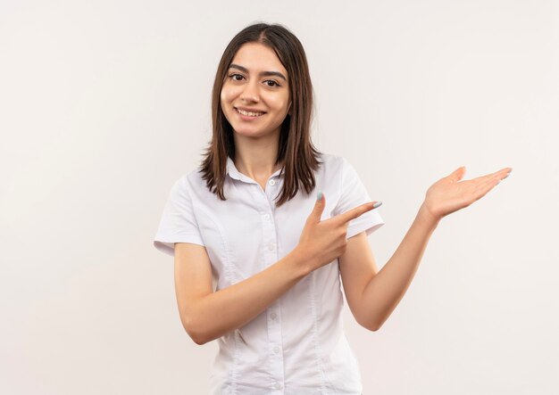 Mujer joven con camisa blanca mirando al frente sonriendo presentando algo con el brazo de su mano apuntando con el dedo hacia el lado de pie sobre la pared blanca