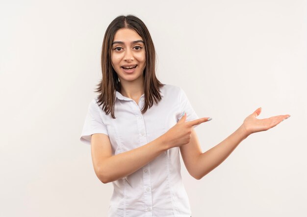 Mujer joven con camisa blanca mirando al frente sonriendo presentando algo con el brazo de su mano apuntando con el dedo hacia el lado de pie sobre la pared blanca