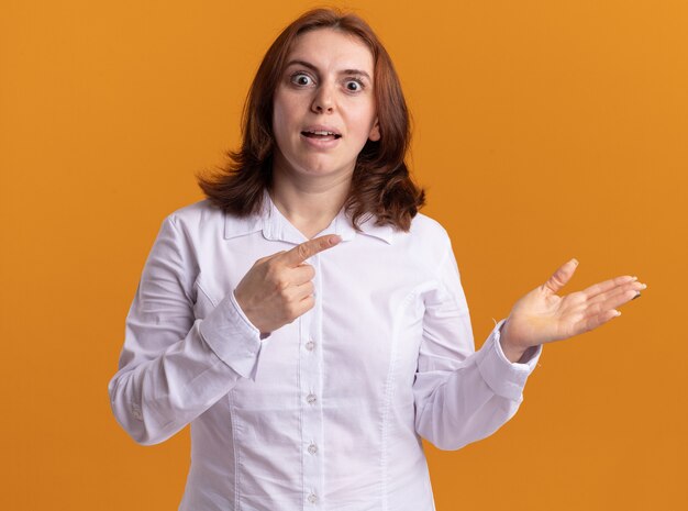 Mujer joven con camisa blanca mirando al frente sorprendido apuntando con el dedo índice hacia el lado de pie sobre la pared naranja