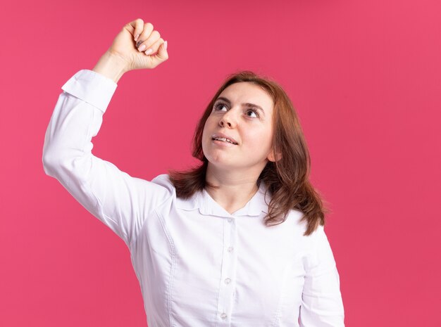 Mujer joven con camisa blanca mirando hacia arriba con expresión de confianza levantando el puño de pie sobre la pared rosa