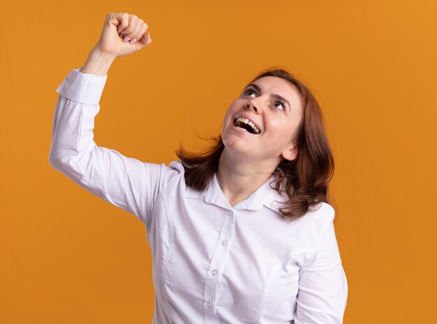 Mujer joven con camisa blanca mirando hacia arriba feliz y emocionado levantando el puño como un ganador de pie sobre la pared naranja