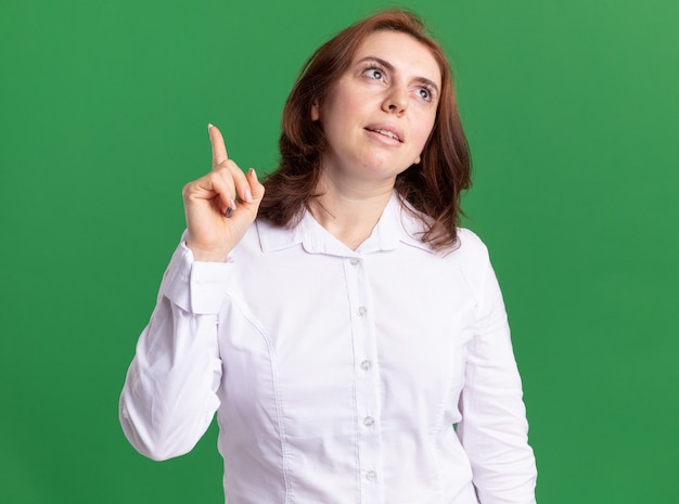 Mujer joven con camisa blanca mirando hacia arriba con una sonrisa en la cara mostrando el dedo índice pensando de pie sobre la pared verde