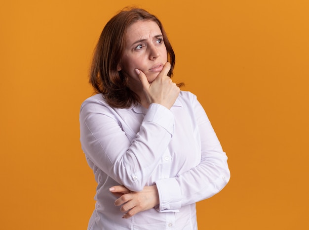 Mujer joven con camisa blanca mirando a un lado con la mano en la barbilla pensando de pie sobre la pared naranja