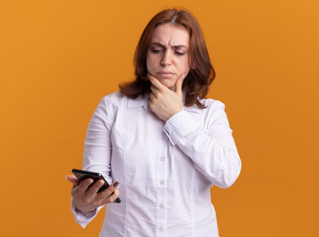 Mujer joven con camisa blanca mirando su teléfono inteligente con expresión pensativa pensando de pie sobre la pared naranja