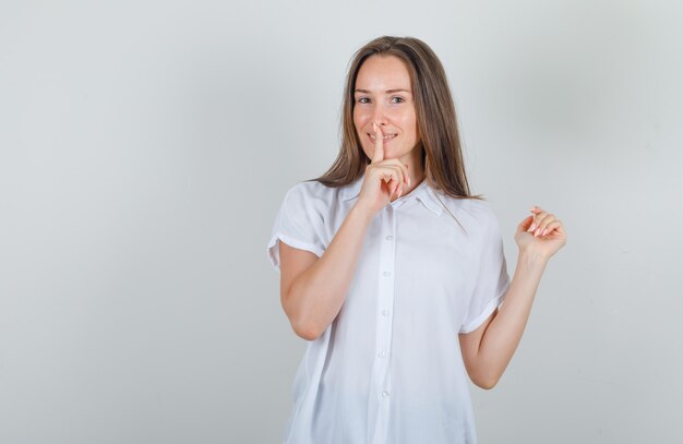 Mujer joven con camisa blanca mostrando gesto de silencio y sonriendo