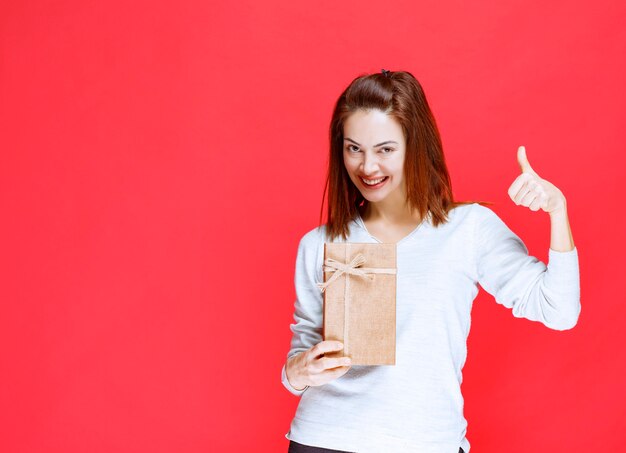 Mujer joven con camisa blanca sosteniendo una caja de regalo de cartón y mostrando un signo de mano positivo