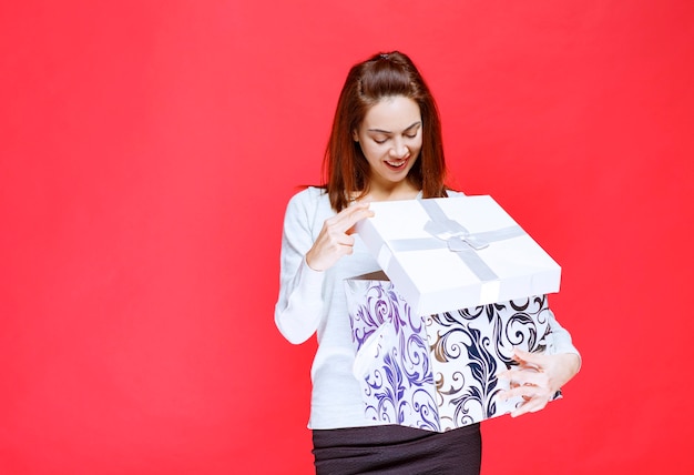 Mujer joven con camisa blanca sosteniendo una caja de regalo impresa, abriéndola y sorprendiéndose