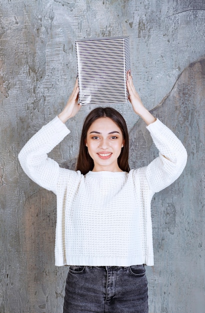 Mujer joven con camisa blanca sosteniendo una caja de regalo plateada sobre la cabeza