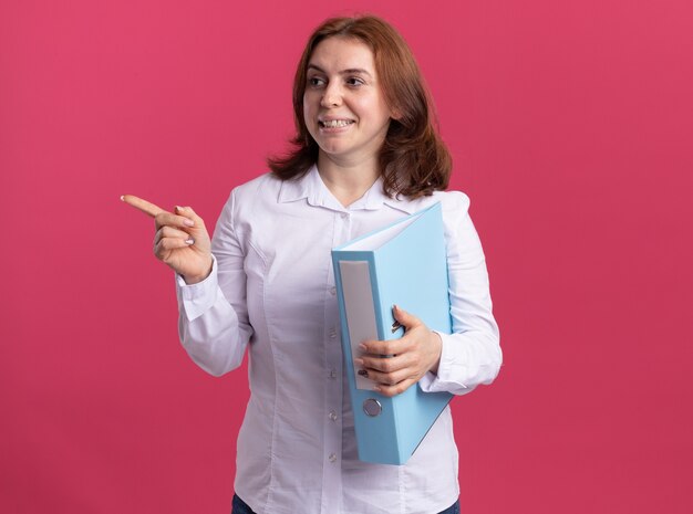 Mujer joven con camisa blanca sosteniendo la carpeta mirando a un lado apuntando con el dedo índice hacia el lado sonriendo alegremente de pie sobre la pared rosa