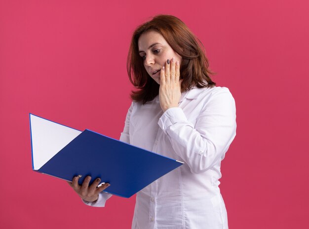 Mujer joven con camisa blanca sosteniendo la carpeta mirándola con expresión confusa de pie sobre la pared rosa