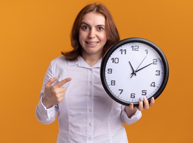 Mujer joven con camisa blanca sosteniendo el reloj de pared mirando al frente con una sonrisa en la cara de pie sobre la pared naranja