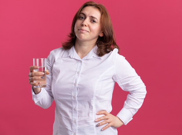 Mujer joven con camisa blanca sosteniendo un vaso de agua mirando al frente sonriendo alegremente de pie sobre la pared rosa