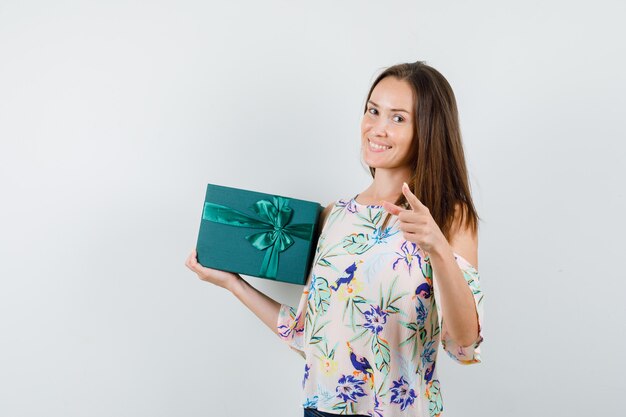 Mujer joven en camisa con caja de regalo y apuntando a la cámara, vista frontal.