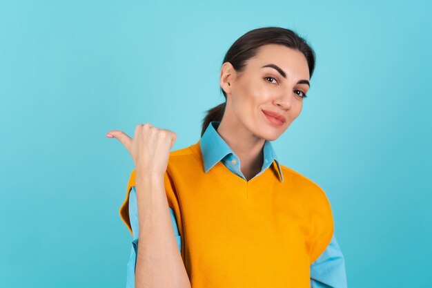Mujer joven con una camisa y un chaleco naranja sobre un fondo turquesa sonriendo alegremente, apuntando con el dedo hacia la izquierda a un espacio vacío
