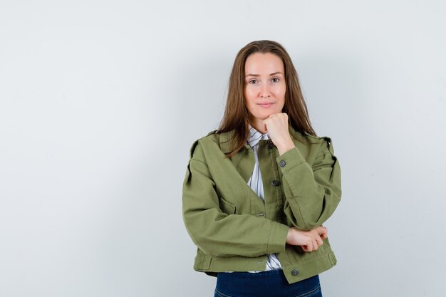 Mujer joven en camisa, chaqueta apoyando la barbilla en el puño y mirando sensible, vista frontal.