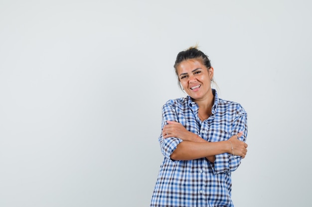 Mujer joven en camisa a cuadros abrazándose a sí misma y mirando adorable.