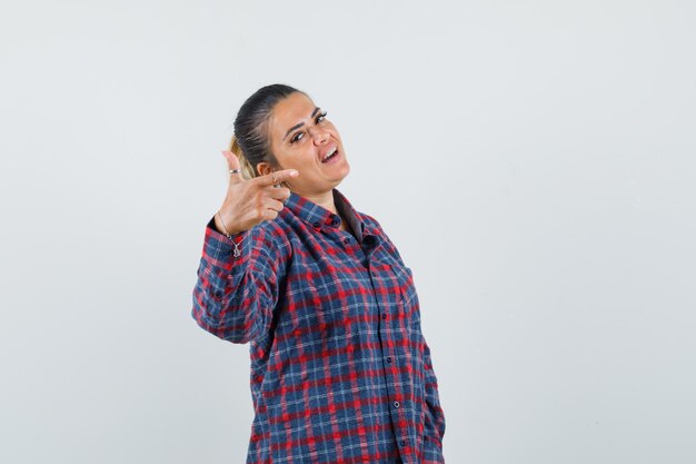 Foto gratuita mujer joven en camisa a cuadros apuntando hacia la derecha con el dedo índice y mirando feliz, vista frontal.
