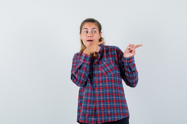 Mujer joven en camisa a cuadros apuntando hacia la derecha con los dedos índices y luciendo bonita