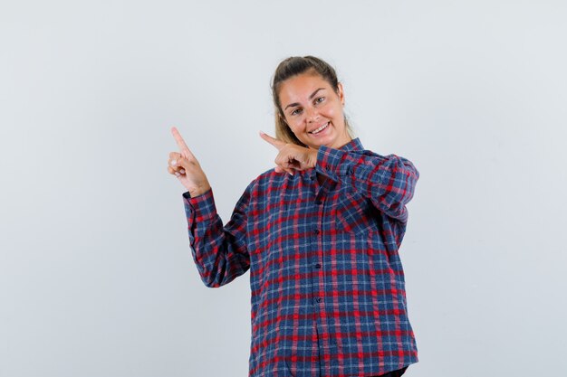 Foto gratuita mujer joven en camisa a cuadros apuntando hacia la parte superior izquierda con los dedos índices y mirando feliz