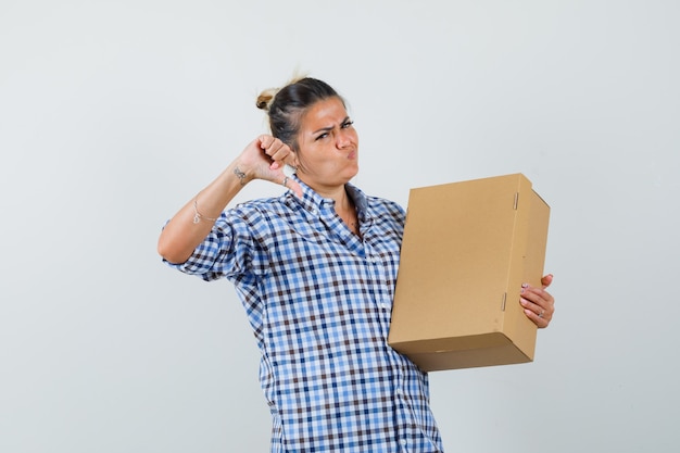 Foto gratuita mujer joven en camisa a cuadros con caja mientras muestra el pulgar hacia abajo y parece disgustado.