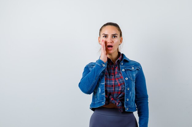 Mujer joven en camisa a cuadros, chaqueta de jean diciendo secreto y mirando sorprendido, vista frontal.