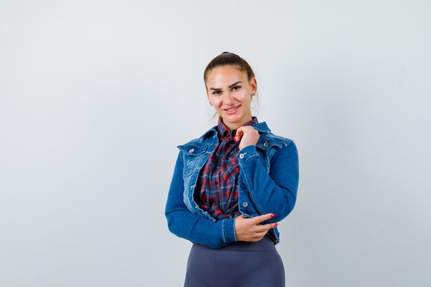 Mujer joven en camisa a cuadros, chaqueta, pantalón posando mientras está de pie y mirando encantador, vista frontal.