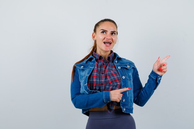 Mujer joven con camisa a cuadros, chaqueta, pantalones apuntando hacia el lado derecho y mirando feliz, vista frontal.