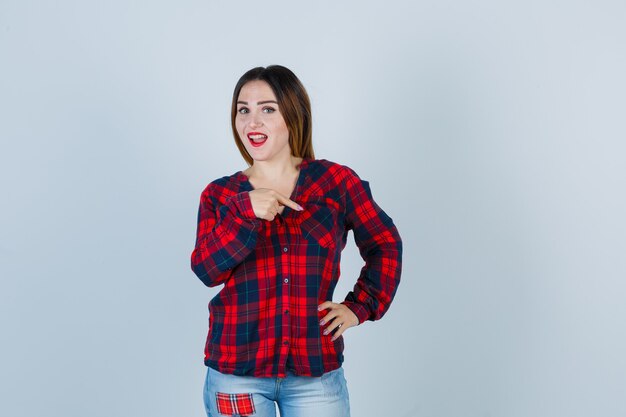 Mujer joven en camisa a cuadros, jeans apuntando hacia abajo con el dedo índice, con la mano en la cintura y mirando alegre, vista frontal.