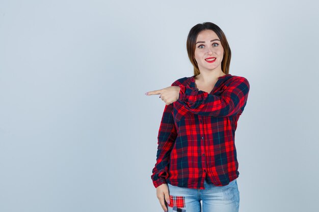 Mujer joven en camisa a cuadros, jeans apuntando hacia la izquierda con el dedo índice y mirando feliz, vista frontal.