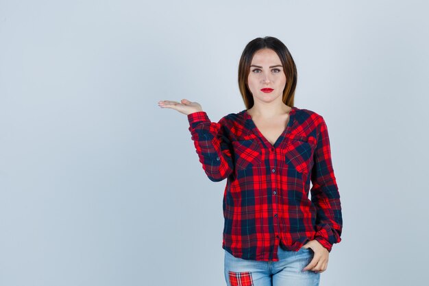 Mujer joven en camisa a cuadros, jeans estirando la mano como sosteniendo algo imaginario, poniendo la mano en el bolsillo y mirando serio, vista frontal.