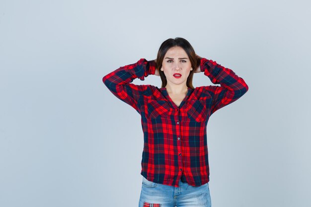 Mujer joven en camisa a cuadros, jeans manteniendo las manos detrás de la cabeza y mirando confundido, vista frontal.