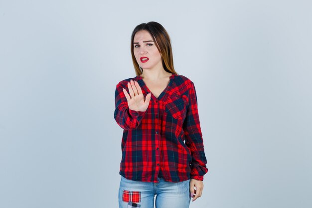 Mujer joven en camisa a cuadros, jeans mostrando señal de stop y mirando acosado, vista frontal.