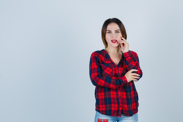 Mujer joven en camisa a cuadros, jeans poniendo la mano cerca de la boca, con la mano en el codo mientras posa y se ve atractiva, vista frontal.