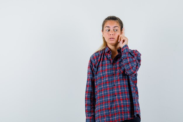 Mujer joven en camisa a cuadros levantando el dedo índice en gesto eureka y mirando pensativo