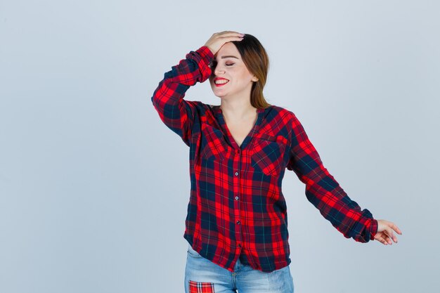 Mujer joven en camisa a cuadros manteniendo la mano en la cabeza y mirando contento, vista frontal.