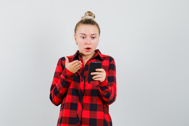 Mujer joven en camisa de cuadros mirando el teléfono móvil y mirando sorprendido