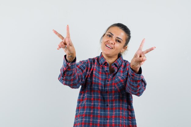 Mujer joven en camisa a cuadros mostrando signos de paz con ambas manos y luciendo bonita vista frontal.