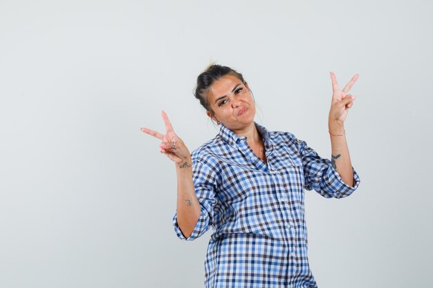 Mujer joven en camisa a cuadros mostrando v-sign y mirando alegre.