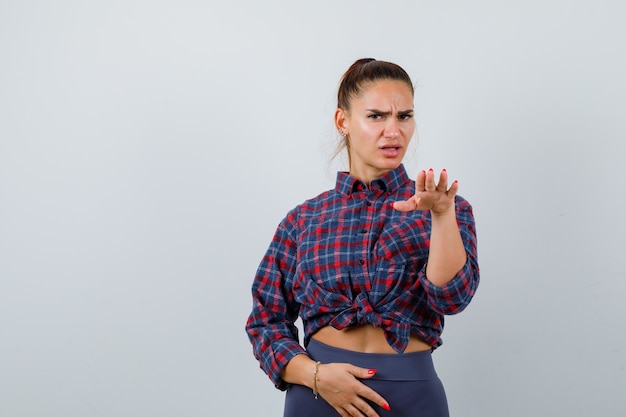 Mujer joven en camisa a cuadros, pantalones haciendo un gesto con la palma hacia afuera para detenerse y mirando seriamente, vista frontal.